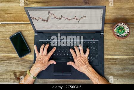 Faltige Hände älterer Frauen, die auf einem Laptop mit Börsencharts arbeiten Stockfoto