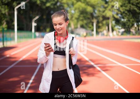 Hübsches Mädchen mit Kopfhörern glücklich mit Handy mit Rucksack auf der Schulter, während die Zeit auf der Rennstrecke des Stadions Stockfoto