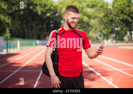 Junger lächelnder Mann in schnurlosen Ohrhörern, der glücklich sein Handy benutzt, während er Zeit im Stadion verbringt Stockfoto