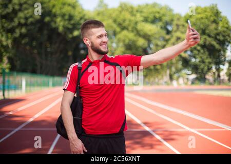 Junger lächelnder Kerl in drahtlosen Kopfhörern glücklich, Fotos auf Frontalkamera des Mobiltelefons im Stadion Stockfoto