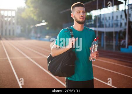 Junger Mann, der überlegt neben sich schaut, mit Sporttasche auf der Schulter und einer Flasche Wasser in der Hand auf der Laufstrecke des Stadions Stockfoto