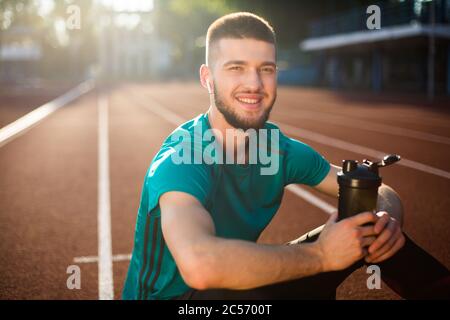 Porträt eines jungen Mannes in drahtlosen Ohrhörern glücklich in der Kamera mit Sportflasche in den Händen, während die Zeit auf der Laufstrecke des Stadions zu verbringen Stockfoto