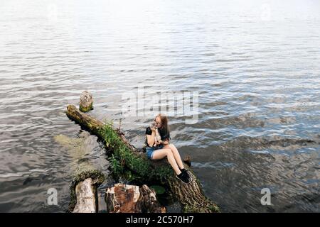 Junge blonde Frau mit blauen Haaren, Brille, Jeans-Shorts und einem schwarzen T-Shirt. Sitzt auf einem schwimmenden Baum in der Nähe des Sees mit einem drei Monate alten Kätzchen Rasse Scottish Straight. Stockfoto