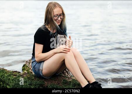 Junge blonde Frau mit blauen Haaren, Brille, Jeans-Shorts und einem schwarzen T-Shirt. Sitzt auf einem schwimmenden Baum in der Nähe des Sees mit einem drei Monate alten Kätzchen Rasse Scottish Straight. Stockfoto