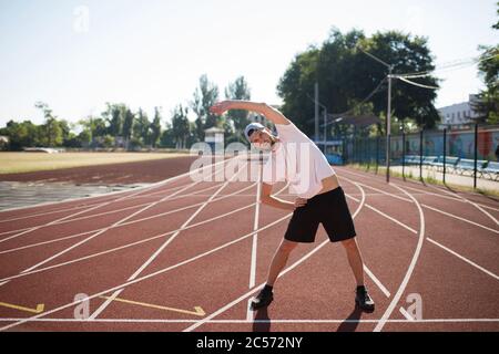 Junge sportliche Kerl in Mütze nachdenklich Blick beiseite beim Stretching auf Laufband des Stadions Stockfoto