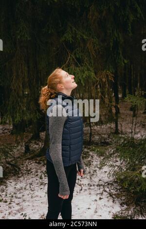 Heitere Rotschopf Frau, die in verschneiten Wäldern aufschaut Stockfoto