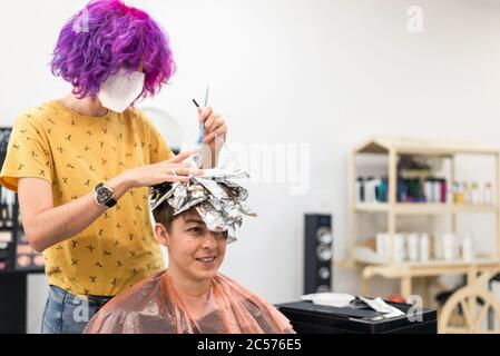 Konzept: Entbindung nach Coronavirus, covid-19. Professioneller Friseur mit Gesichtsmaske und Handschuhen. Blondine Highlights mit Aluminiumfolie putzen Stockfoto