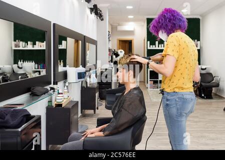 Konzept: Entbindung nach Coronavirus, covid-19. Details. Professioneller Friseur mit lila Haaren. Trocknen des Kopfes mit einer Maschine zu einem lächelnden wom Stockfoto