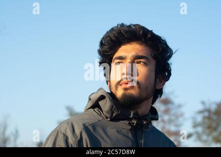Ein hübscher junger Mann schaut abends von der Kamera weg. Wenige Bäume und ein klarer blauer Himmel im Hintergrund. Mann trägt eine Sportjacke und hat Stockfoto