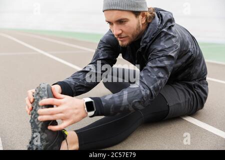 Zugeschnittenes Bild eines motivierten jungen Sportlers, der auf der Stadionrennbahn arbeitet und Stretching-Übungen macht Stockfoto