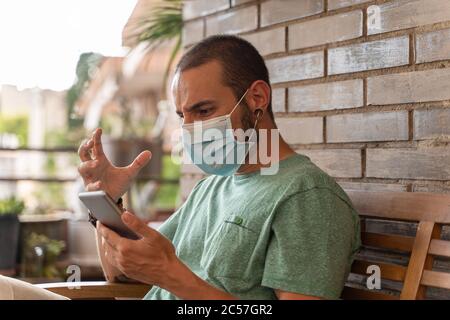 Junger Mann mit einem Bart durch eine Maske geschützt macht Videokonferenz mit seinem Handy auf einer Terrasse. Der Mann ist nervös und wütend. Business und Sicherheit c Stockfoto
