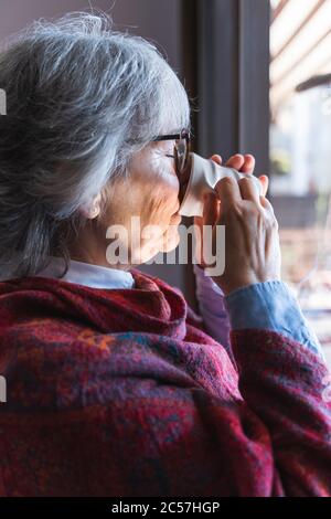 Vertikale Aufnahme einer älteren Frau mit Gläsern, die Tee aus einer weißen Keramikschale neben einem Fenster trinken Stockfoto