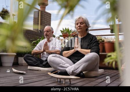 Seniorenpaar meditiert auf der Holzterrasse, umgeben von Pflanzen, mit einer Nahaufnahme von einigen unfokussierten Pflanzen, Kerzen und Weihrauch Stockfoto