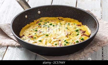 Omelette mit Prosciutto Erbsen und Kräutern in Keramikpfanne auf Tisch Stockfoto