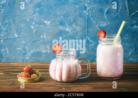 Reife Erdbeeren im Deckel aus einer Dose. Im Glas frischer Erdbeer Smoothie auf einem Holztisch. Blauer Hintergrund Stockfoto