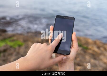 Nahaufnahme einer Frau, die das Mobiltelefon hält und die andere den Bildschirm berührt, wobei der Hintergrund von Felsen und das Meer unscharf ist Stockfoto