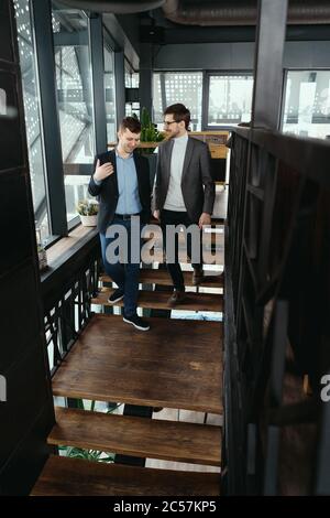 Zwei Männer, die auf Holztreppen hinunterlaufen, plaudern drinnen. Kommunikationskonzept Stockfoto