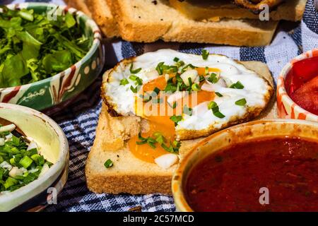 Rustikale Komposition mit Würstchenbrötchen, Spiegelei auf Toast, verschiedenen Schüsseln mit Sauce und gehacktem Gemüse. Stockfoto