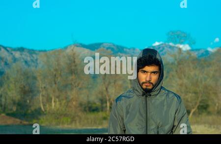 Ein hübscher junger Mann mit Kapuzenpullover und Blick in die Kamera mit strengen Gesichtsausdrücken am Abend. Bäume, klarer blauer Himmel und Berge in Stockfoto