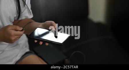 Die Hände einer Frau laden ein Smartphone auf, während sie auf dem schwarzen Ledersofa sitzt. Stockfoto