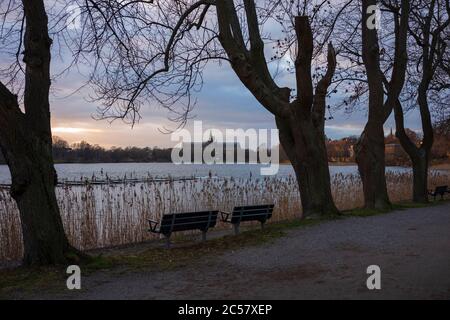 Leere Parkbänke und blattlose Bäume entlang der Wasserstraße Drugardsbrunnsviken bei einem Winteruntergang in Stockholm, Schweden. Stockfoto