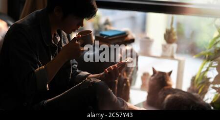 Ein intelligenter Mann trinkt heißen Kaffee, während mit einem Smartphone in der Hand und sitzt auf Ledercouch neben seiner schönen Katze über komfortablen Wohnzimmer als Stockfoto