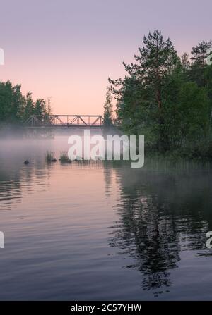 Neblige Sommerlandschaft und alte Eisenbrücke mit schönen ruhigen See in der Sommernacht in Finnland Stockfoto