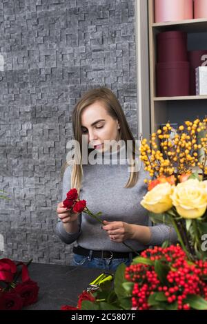 Hübsche Frau Floristin wählt Rosenblätter aus. Die Blondine sammelt einen Blumenstrauß aus perfekten roten Rosen. Stockfoto