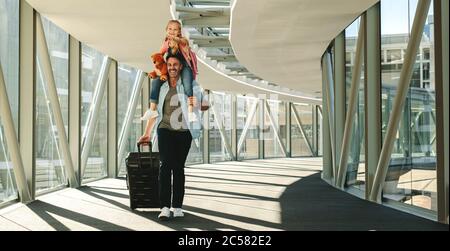Mädchen auf der Schulter ihres Vaters am Flughafen sitzen. Vater und Tochter mit Gepäck gehen durch einen Tunnel, um ihren Flug zu besteigen. Stockfoto