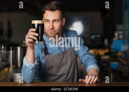 Brauer und Barkeeper. Ein gutaussehender, stilvoller Mann in der Schürze schaut auf ein Glas Bier Stockfoto