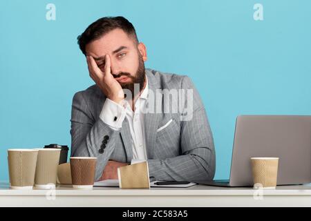 Müde Geschäftsmann umgeben von leeren Kaffeetassen sitzen an seinem Schreibtisch mit Laptop-Computer auf blauem Hintergrund Stockfoto