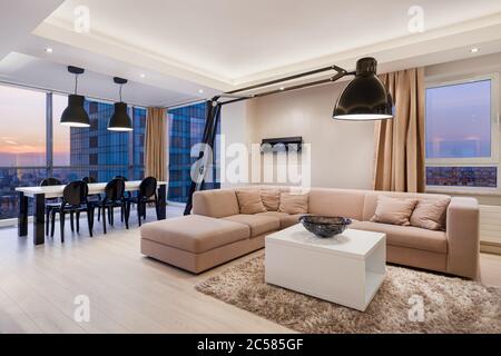 Elegante Wohnung mit beigem Wohnzimmer mit großer Lampe und stilvollem Essbereich mit Blick auf den Sonnenuntergang Stockfoto