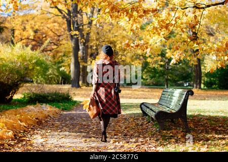 Elegante Rothaarige Frau in karierten Mantel und schwarze Baskenmütze geht durch den Park, hält trockene Ahornblätter und Rucksack an sonnigen Tag, Rückansicht.Herbststimmung Stockfoto