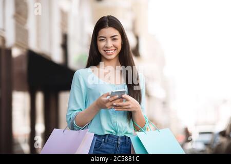 Glücklich Asiatische Shopper Mädchen Posiert Im Freien Mit Smartphone Und Einkaufstaschen Stockfoto