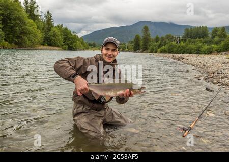 Ein Fischer hält einen männlichen Buckellachs in der Hand, während er in einem Fluss watet, mit Wald und Bergen im Hintergrund. Stockfoto