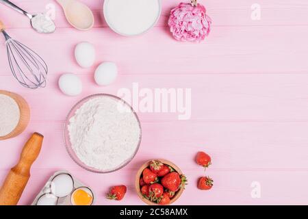 Zutaten zum Backen köstlicher SommerErdbeerkuchen, frische Beeren, Eier, Mehl und Zucker neben Schneebesen und Nudelholz auf rosafarbenem Tisch mit Copy Spa Stockfoto