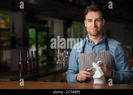 Moderne Arbeit des Barkeepers. Schöner Mann in Schürze mit stilvollen Haarschnitt, reibt Glas Stockfoto