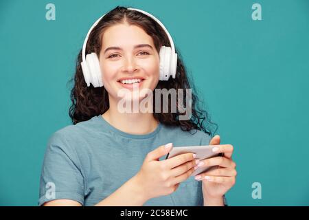 Porträt von fröhlichen jungen Frau in kabellosen Kopfhörern Filme auf dem Smartphone vor blauem Hintergrund Stockfoto
