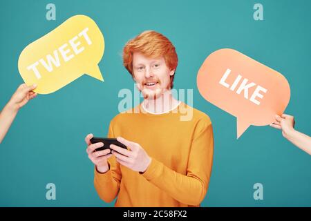 Portrait von fröhlichen jungen Rotschopf Mann in orange Pullover süchtig nach sozialen Medien mit Smartphone, Hände erhöhen Tweet und wie Zeichen Stockfoto