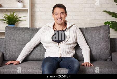 Glücklicher Mann in einem weißen Pullover sitzt auf einem Sofa zu Hause Stockfoto