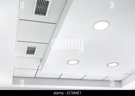 Mehrstufige Decke mit dreidimensionalen Aussprüngen und einer abgehängten Fliesendecke mit eingebautem runden LED-Licht und Kühlergrill-Lüftungssystem. Stockfoto