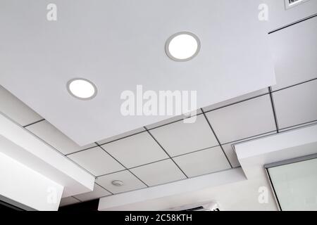 Mehrstufige Decke mit dreidimensionalen Aussprüngen und einer abgehängten Fliesendecke mit eingebautem runden LED-Licht, weiße Farbe. Stockfoto