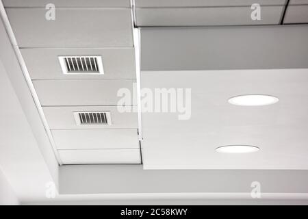 Mehrstufige Decke mit dreidimensionalen Aussprüngen und einer abgehängten Fliesendecke mit eingebautem runden LED-Licht und einem Lüftungsgitter. Stockfoto