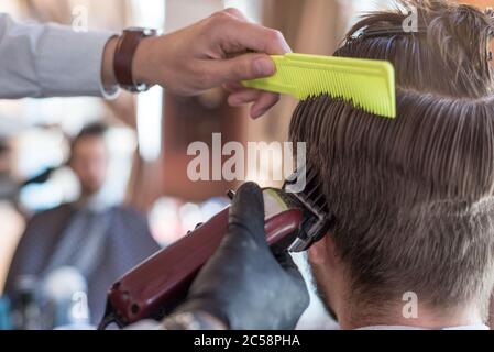 Ein Friseur schneidet einen bärtigen jungen Kerl mit einem Haarschneider und kämmt die Haare auf seinem Kopf. Die Arbeit des Meisters im Männerhaarschnitt in einem Friseurladen Stockfoto