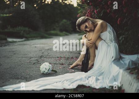 Einsame Braut am Hochzeitstag geworfen, Alkohol aus einer Flasche trinken, sitzen auf dem Bürgersteig in einem weißen Kleid. Vor dem Hintergrund der roten Rose Stockfoto