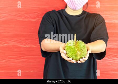 Junge Frau trägt eine Schutzmaske, um eine Infektion des Corona-Virus (Covid-19) in einem schwarzen Hemd mit einem reifen Durian zu verhindern, bereit, mit dem roten w zu essen Stockfoto