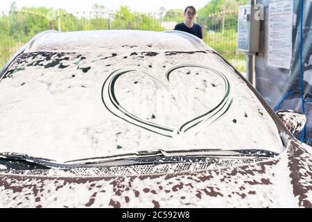 Ein Herz wird auf dem Autoglas in Schaum bei einer Autowäsche gezeichnet. Der Mann im Hintergrund. Symbol des Respekts für das Auto des Kunden Stockfoto