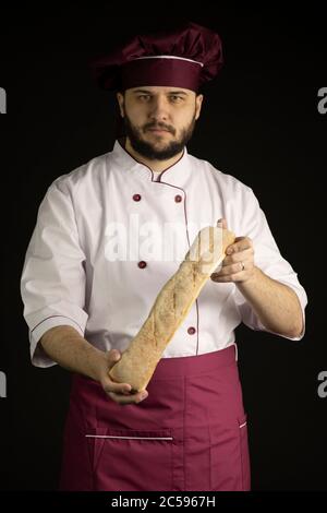 Gutaussehender bärtiger Chefkoch in Uniform hält Baguette, Laib französisches Brot Stockfoto