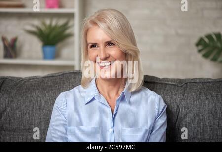 Glückliche ältere Frau, die sich zu Hause auf dem Sofa entspannt. Nahaufnahme eines reifen blonden Frau lächeln und wegschauen. Stockfoto