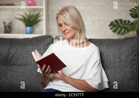 Schöne Frau mittleren Alters liest ein Buch, während sie zu Hause auf einem Sofa sitzt Stockfoto